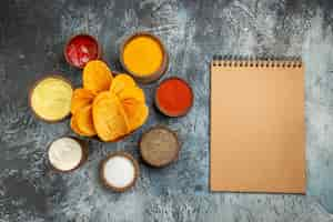 Free photo horizontal view of crispy chips on wooden cutting board served with different spices mayonnaise ketchup and notebook on gray table