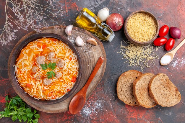 Horizontal view of delicious noodle soup with chicken on wooden cutting board greens spoon garlic tomato and bread slices on dark background