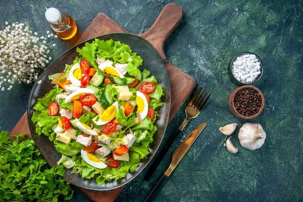 Horizontal view of delicious salad with many fresh ingredients on wooden cutting board spices oil bottle garlics cutlery set on black green mix colors background