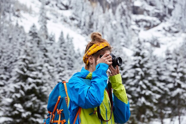 Horizontal view of female hiker poses in snowy mountains, reaches top, makes photo with camera from hill