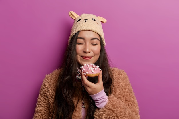 Horizontal view of lovely woman with pleased expression, closes eyes and smells delicious doughnut, wears winter clothing, isolated over lilac wall