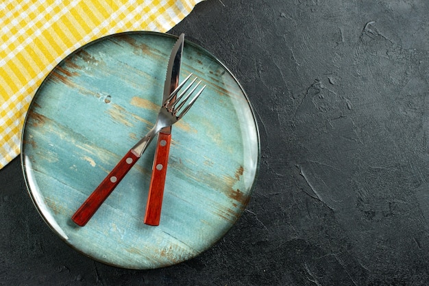 Horizontal view of meal cutlery in cross on a blue plate and yellow stripped towel on the right side on dark surface