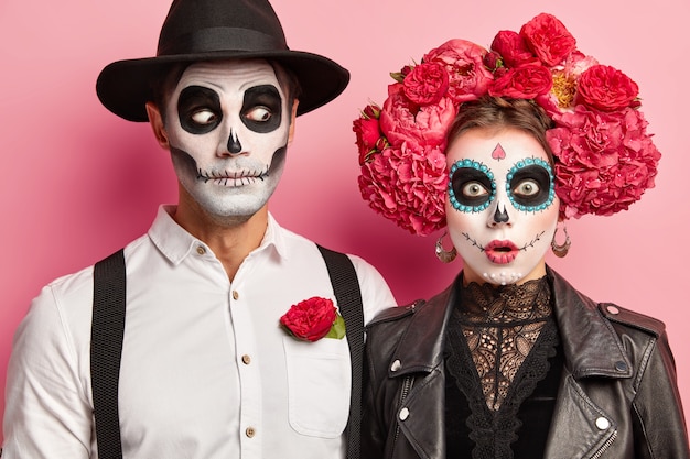 Horizontal view of surprised couple with scary face, painted sugar skulls and smiles, celebrate popular carnival together