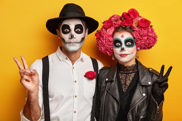 Horizontal view of woman and man wear bright makeup, make peace gesture, wear traditional clothes, celebrate Dead of Death, isolated over yellow background.