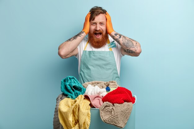 Horrified ginger young man keeps hands on head, opens mouth widely