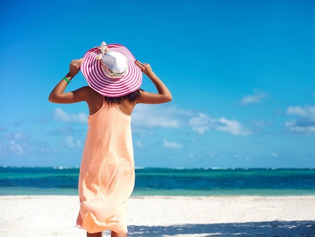 Hot beautiful woman in colorful sunhat and dress walking near beach ocean on hot summer day on white sand