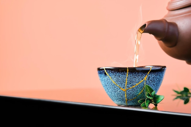 Free photo hot green tea is poured from a ceramic teapot into a bowl. selective focus on the blue cup. steam will rise above the mug reclaimed ceramic blue cup, second life of things, recycling or kintsugi