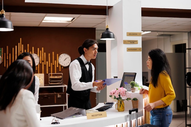 Hotel guest receiving registration form from helpful asian male concierge. Relaxed asian middle aged woman on vacation making necessary preparation for check in at modern resort