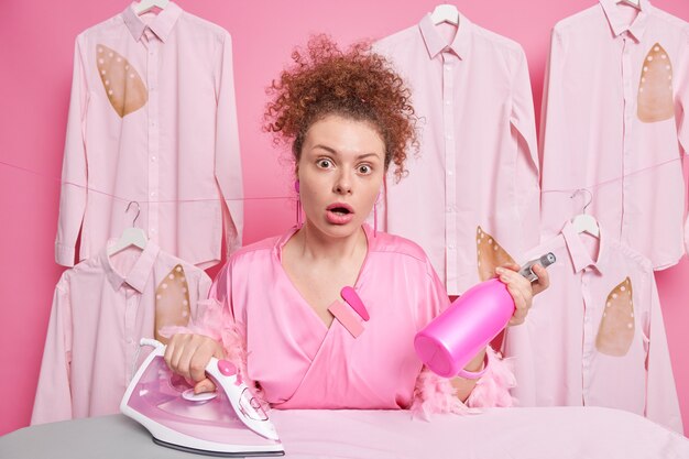 Household and ironing concept. Surprised stunned curly housewife holds detergent spray irons family clothes keeps mouth opened busy doing daily domestic routines dressed in pink dressing gown