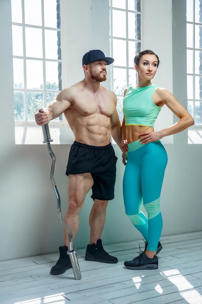 Huge shirtless bearded bodybuilder with sporty woman in azure sportswear posing in natural light near window.