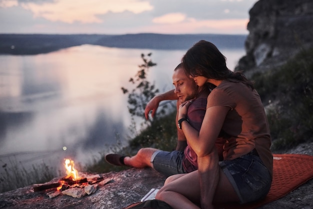 Free photo hugging couple with backpack sitting near the fire on top of mountain enjoying view coast a river or lake.