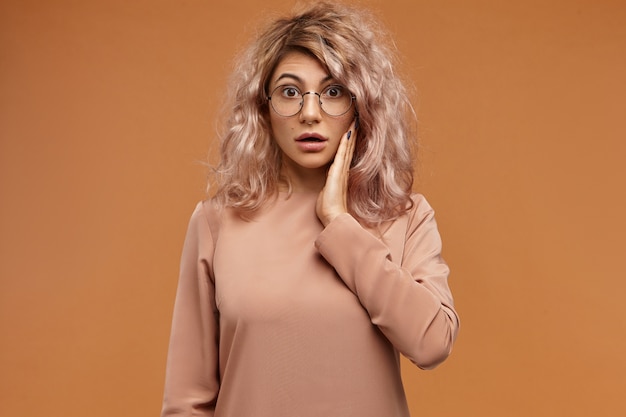 Free photo human emotions, reaction and feelings.  shot of emotional funny hipster girl in round eyeglasses, having puzzled look, holding cheek and opening mouth