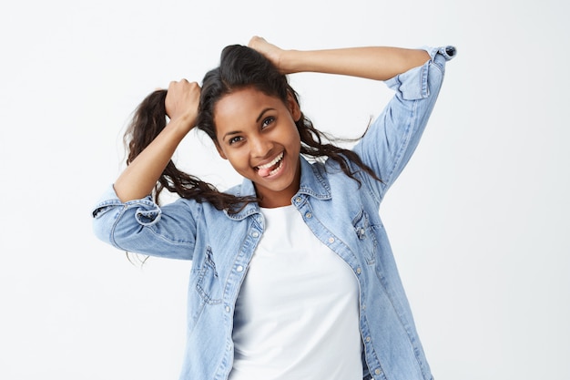 Human face expressions and emotions. Portrait of young playful African-American woman in denim shirt playing with her hair, holding her hair with both hands, with mouth open in joyful smile.