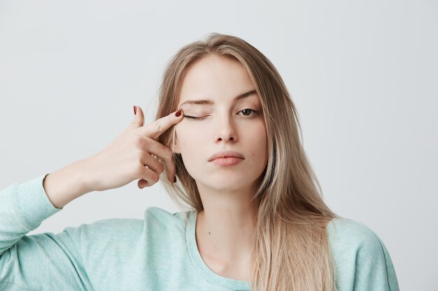 Human face expressions.Thoughtful young beautiful female with blonde dyed straight hair in light blue long-sleeved sweater touching with finger eye