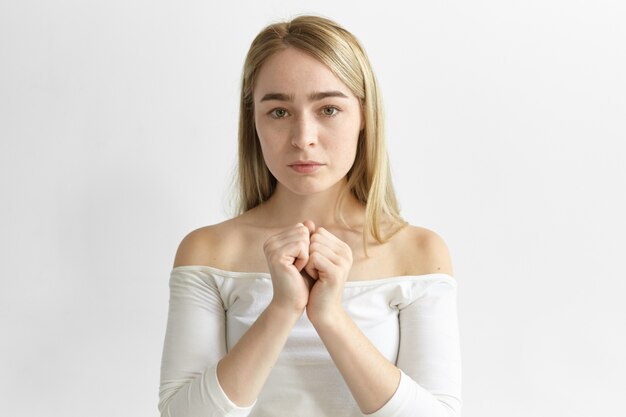 Human facial expressions and body language. Picture of stylish young Caucasian female with loose blonde hair posing indoors, holding two clenched fists together, having uneasy deep in thoughts look