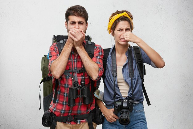 Human facial expressions, emotions and feelings. Tourism and travel. Active young couple in tourist clothing, carrying backpacks, binocular and photo camera pinching noses because of disgusted stink