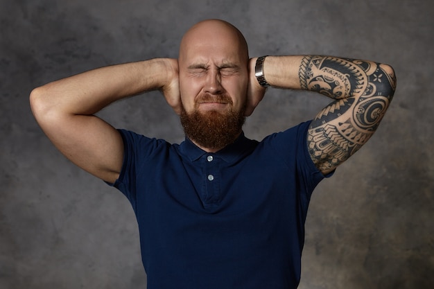 Human facial expressions. Horizontal shot of frustrated irritated stylish young tattooed male with bushy beard keeping eyes closed and covering ears having painful look, stressed with loud noise