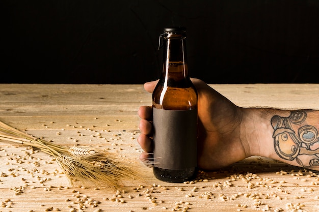 Free photo human hand holding alcoholic bottle with ears of wheat on wooden surface