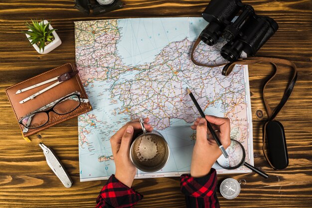 Human hand with cup of coffee marking destinations on map