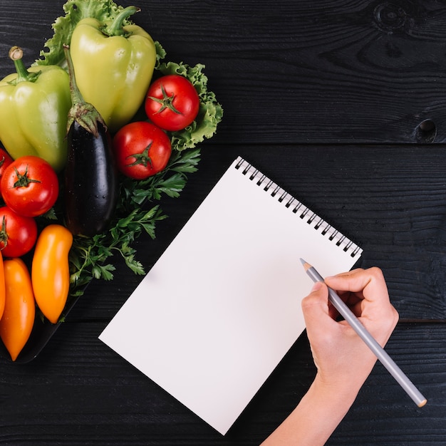 Free photo human hand writing on spiral notepad with fresh vegetables on black wooden backdrop