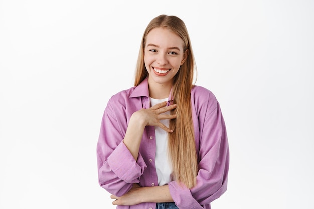 Humble cute blond girl looking touched and flattered smiling pleased thanking for something standing in stylish purple shirt against white background