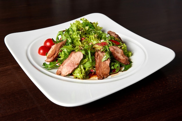 Hungry Closeup of a sliced grilled steak served with green leaves salad and cherry tomatoes on a white square plate on the table at the restaurant .