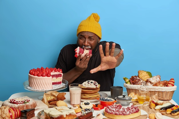 Hungry displeased plump Afro man keeps palm forward to camera, bites huge piece of creamy cake, gets much calories, surrounded with tasty baked products