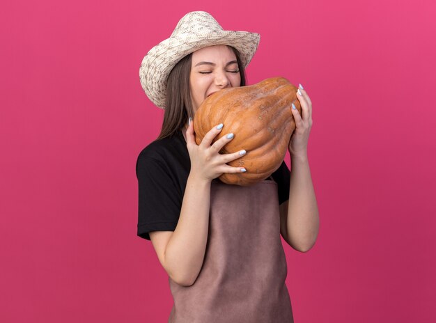 Hungry pretty caucasian female gardener wearing gardening hat pretending to bite pumpkin isolated on pink wall with copy space