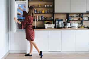 Free photo hungry woman looking for food in the fridge at home but dont have much there white kitchen furniture home wear red silk robe