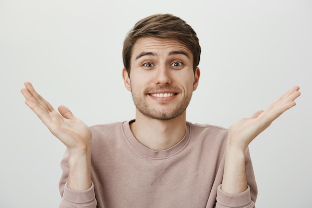 Husband has no idea Indoor portrait of beautiful young man with bristle shrugging and raising palms smiling awkwardly as if apologizing standing against gray background confused