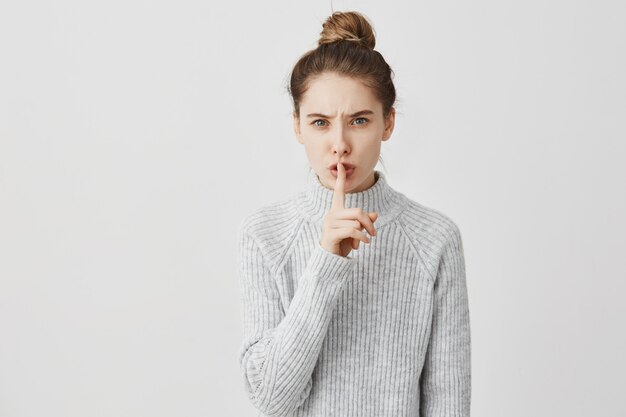 Hush! Headshot of caucasian woman holding index finger on lips. Female receptionist with dark hair tied in bun asking to keep quiet saying shh over white wall. Silence concept