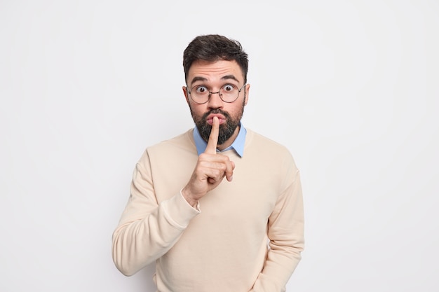 Hush do not say that. Surprised bearded man presses index finger to lips asks not to spread false rumors stares shocked, makes taboo gesture wears spectacles and sweater poses indoor