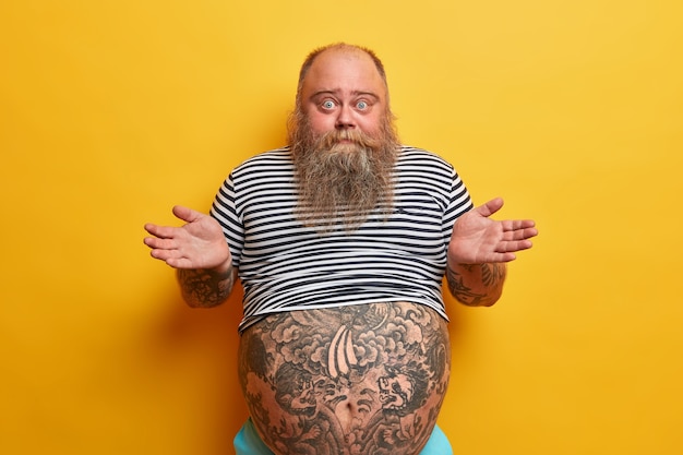 Free photo i dont care about rules. indifferent careless questioned bearded man shrugs shouldres with hesitation, puzzled by stupid question, has very big tummy, wears striped t shirt, no idea how it happened