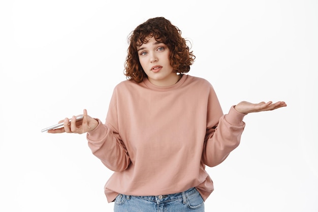 Free photo i dont know. confused young beautiful woman shrugging shoulders, holding smartphone and looking clueless, have no idea, dont understand how mobile app works, standing over white background