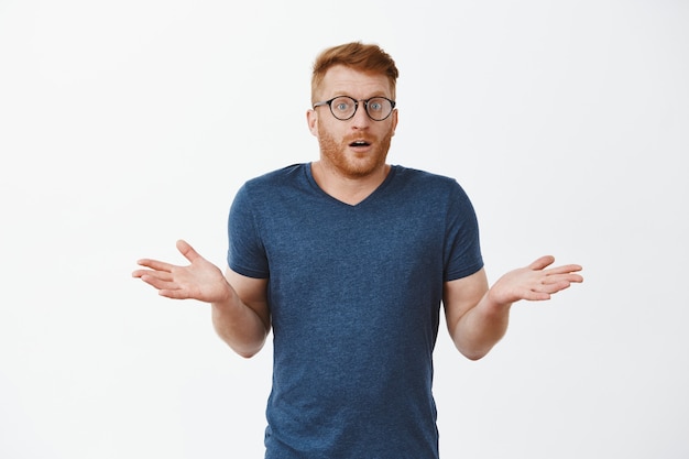 Free photo i have no idea how that happened. portrait of clueless cute masculine male with red hair in glasses and t-shirt, shrugging with spread palms and gazing confused and uncertain over grey wall