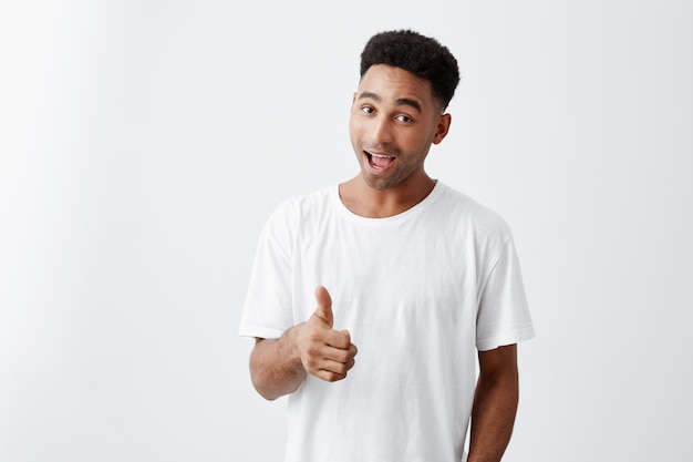 I like it. Young Attractive dark-skinned male university student with afro hairstyle in casual white t-shirt smiling, showing thumb up in camera with happy and excited expression