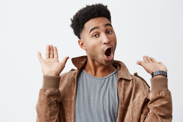 I'm out of this. Portrait of young funny dark-skinned african male with curly hair in grey t-shirt under brown jacket being shocked hearing provoking news about old friend.