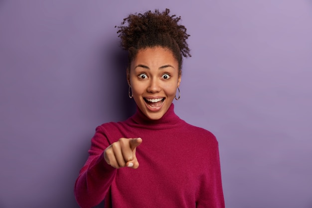 I need you. Happy curly teenage girl indicates  with fore finger, picks candidate, has positive surprised expression, laughs out loud, poses over purple wall, says incredible.