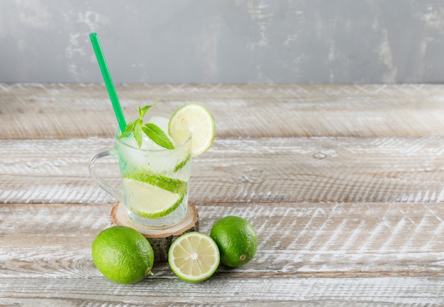 Free photo icy mojito cocktail with limes, mint straw in a cup on wooden and plaster background, high angle view.