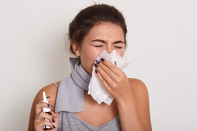 ill allergic woman blowing running nose, having got flu or catch cold, sneezing in handkerchief, posing with closed eyes isolated on white, holding nasal spray in hand.