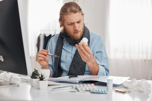 Ill bearded male office worker in blue shirt and scarf with spectacles concentrated on reading prescription of pills. Young manager with flu, sits at workplace surrounded by drugs, tablets, vitamins