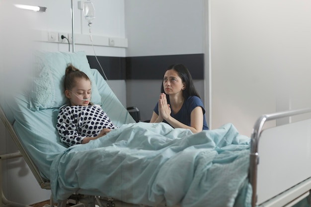 Free photo ill kid under treatment sleeping in hospital pediatrics ward room while worried mother sitting next to her. sick little girl resting on patient bed while mother praying beside her