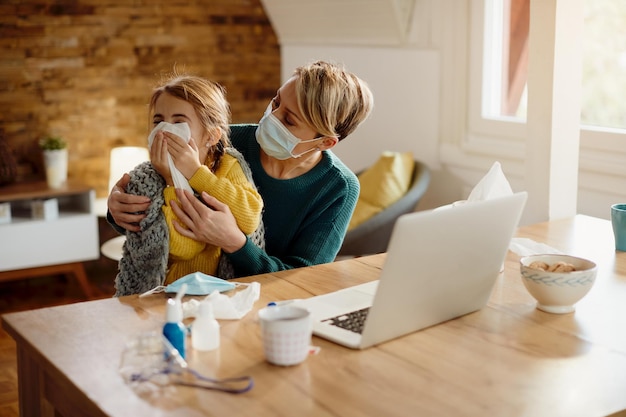 Ill little girl sneezing into a tissue while sitting on mother's lap at home