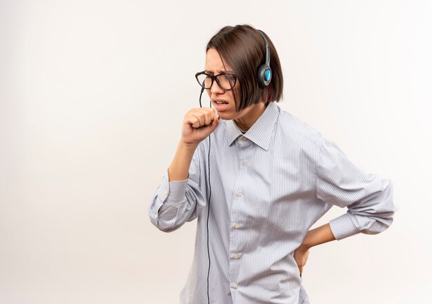 Ill young call center girl wearing glasses and headset keeping fist near mouth coughing and putting hand on waist isolated on white  with copy space