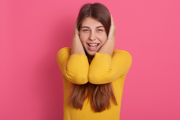 Free photo image of active funny adorable young female opening mouth widely, covering her ears with hands, screaming, avoiding loud noise, wearing yellow sweatshirt. emotions concept.