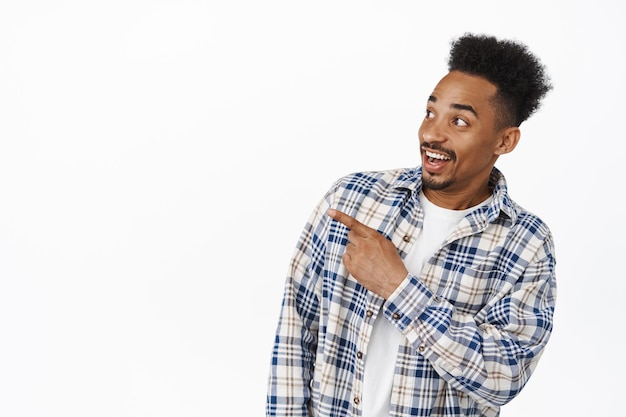 Image of african american young man look surprised, pointing and looking left at sale advertisement with happy amazed smile, checking out promo deal, white background.