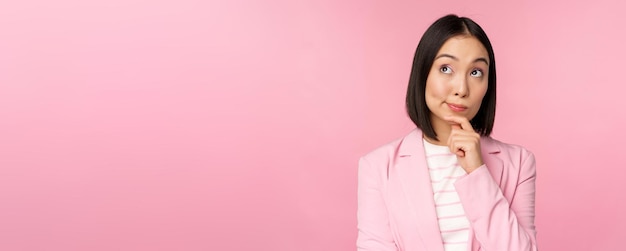 Free photo image of asian businesswoman standing in thinking pose brainstoming wearing suit korean saleswoman entrepreneur posing against pink background