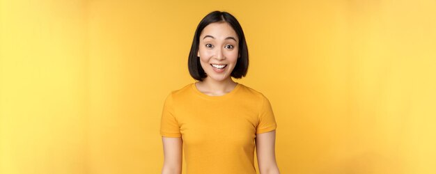 Image of asian girl looking surprised reacting amazed raising eyebrows impressed standing over yellow background