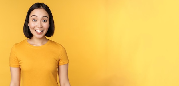 Image of asian girl looking surprised reacting amazed raising eyebrows impressed standing over yellow background