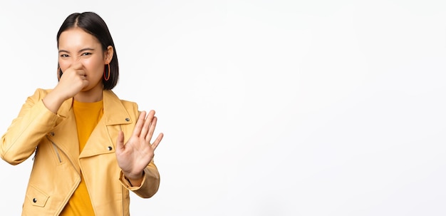 Image of asian girl shuts nose and reject smth with disgusting bad smell shuts nose standing over white background